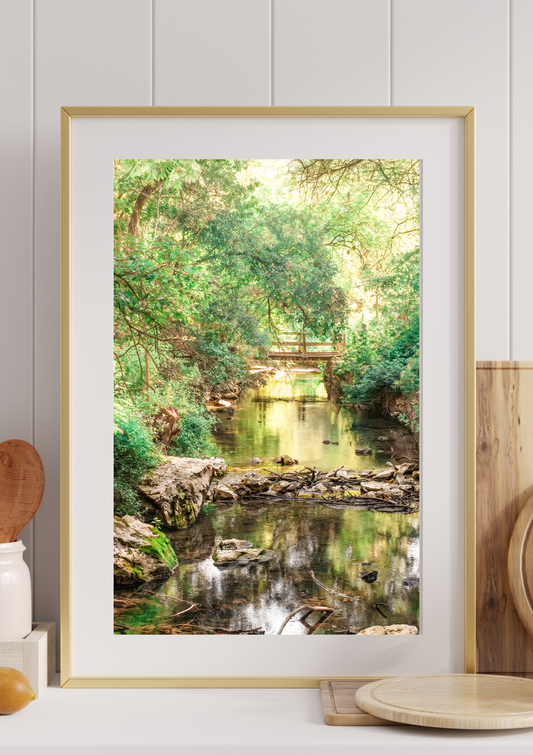 Framed wall art mockup of Slow Current by Preiss Photography displayed in a cozy office setting, featuring a peaceful creek scene with green trees, soft reflected light, and a small wooden bridge in the background.