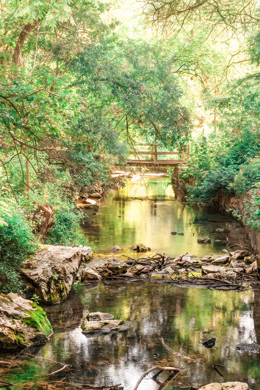 Peaceful Texas Hill Country creek scene with slow-moving water, soft reflections, rocks, lush green trees, and a small wooden bridge in the distance.