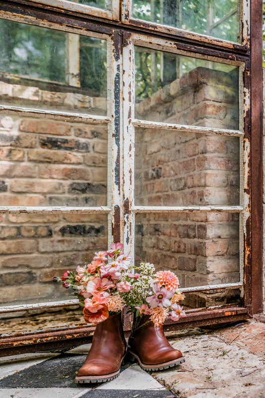 Leather boots filled with pink and peach flowers in front of a rustic brick wall and weathered window.