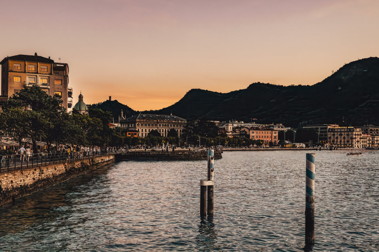 Evening view of Lake Como, Italy, with calm water, warm dusk sky, shoreline buildings, and dark hills in the background.