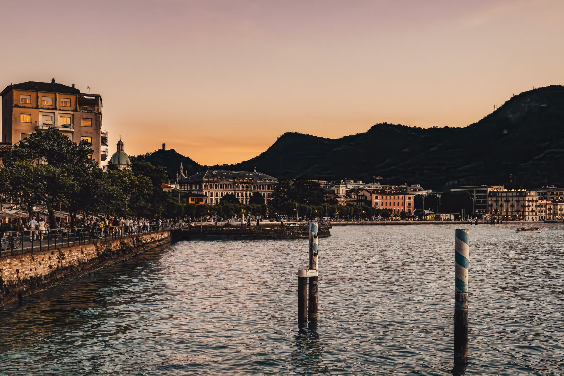 Evening view of Lake Como, Italy, with calm water, warm dusk sky, shoreline buildings, and dark hills in the background.