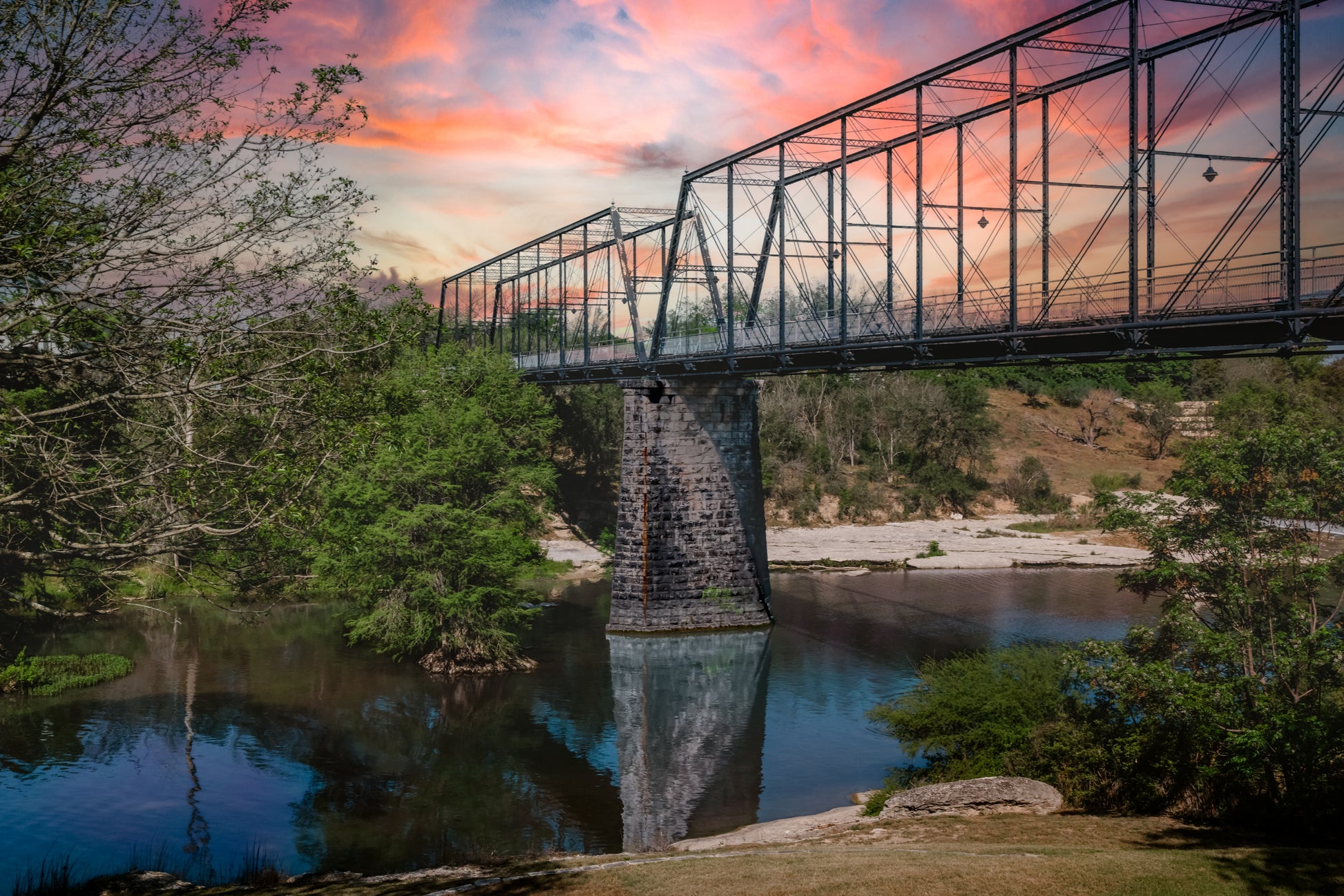 Historic Faust Street Bridge over the Guadalupe River in New Braunfels, Texas, with colorful sunset clouds and river reflections.