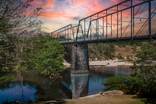 Historic Faust Street Bridge over the Guadalupe River in New Braunfels, Texas, with colorful sunset clouds and river reflections.