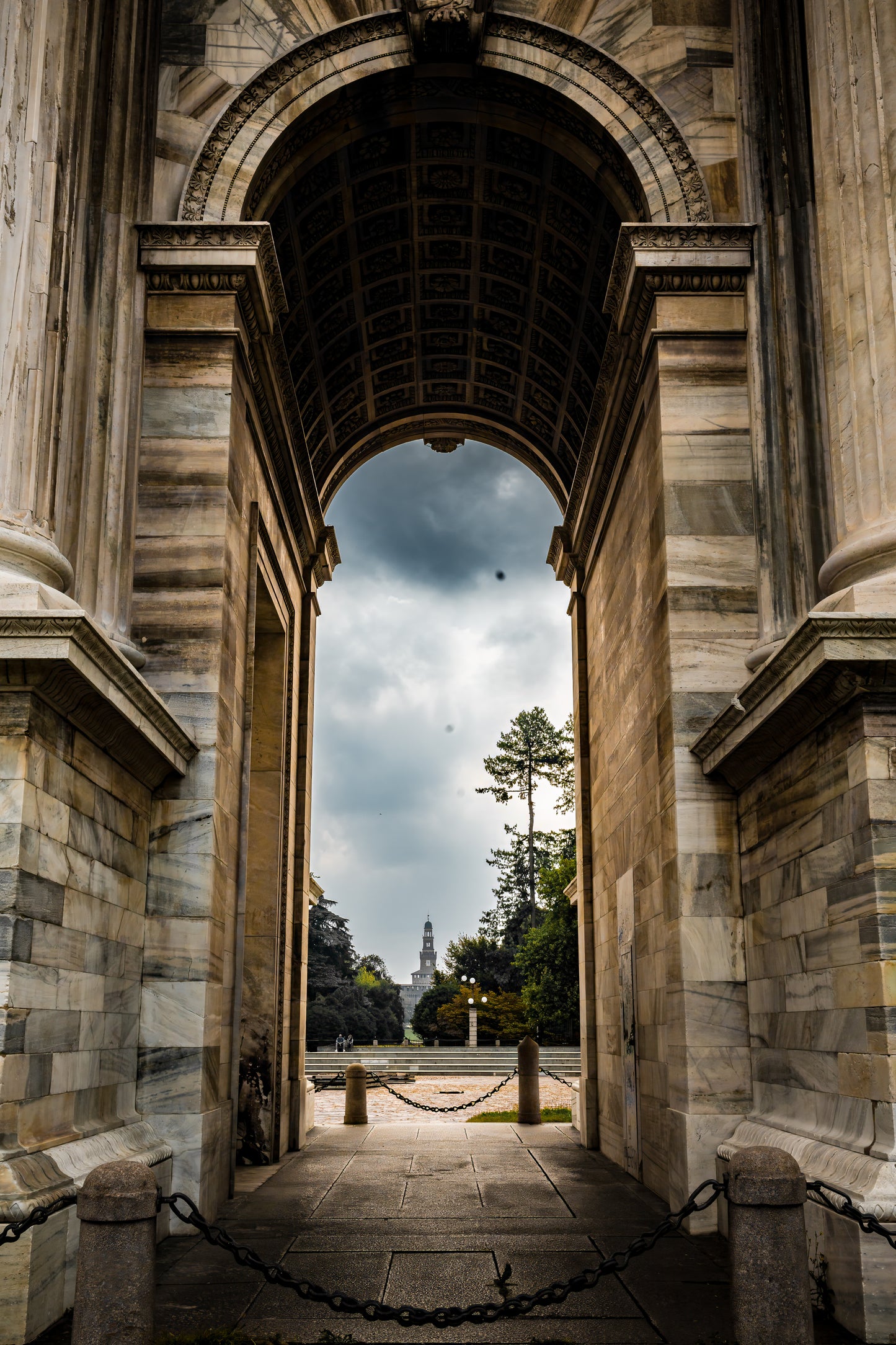 View through the historic Arco della Pace in Milan, Italy, with grand marble architecture, cloudy sky, and a distant city landmark beyond.