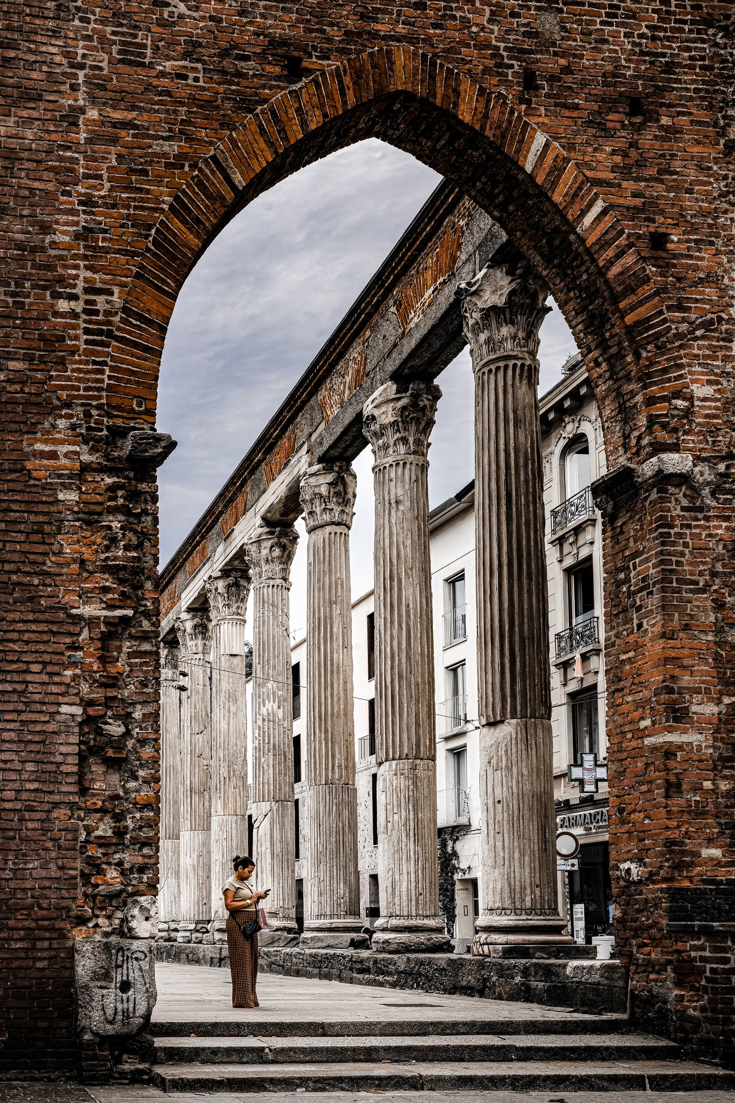 Historic Columns of San Lorenzo in Milan, Italy, framed by a brick arch with ancient Corinthian columns and layered city architecture.