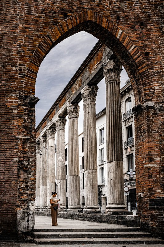 Historic Columns of San Lorenzo in Milan, Italy, framed by a brick arch with ancient Corinthian columns and layered city architecture.
