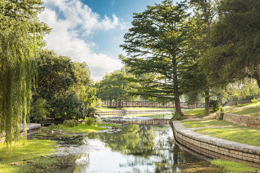Morning light over Landa Park in New Braunfels, Texas, with cypress trees, water reflections, stone pathways, and a bridge.