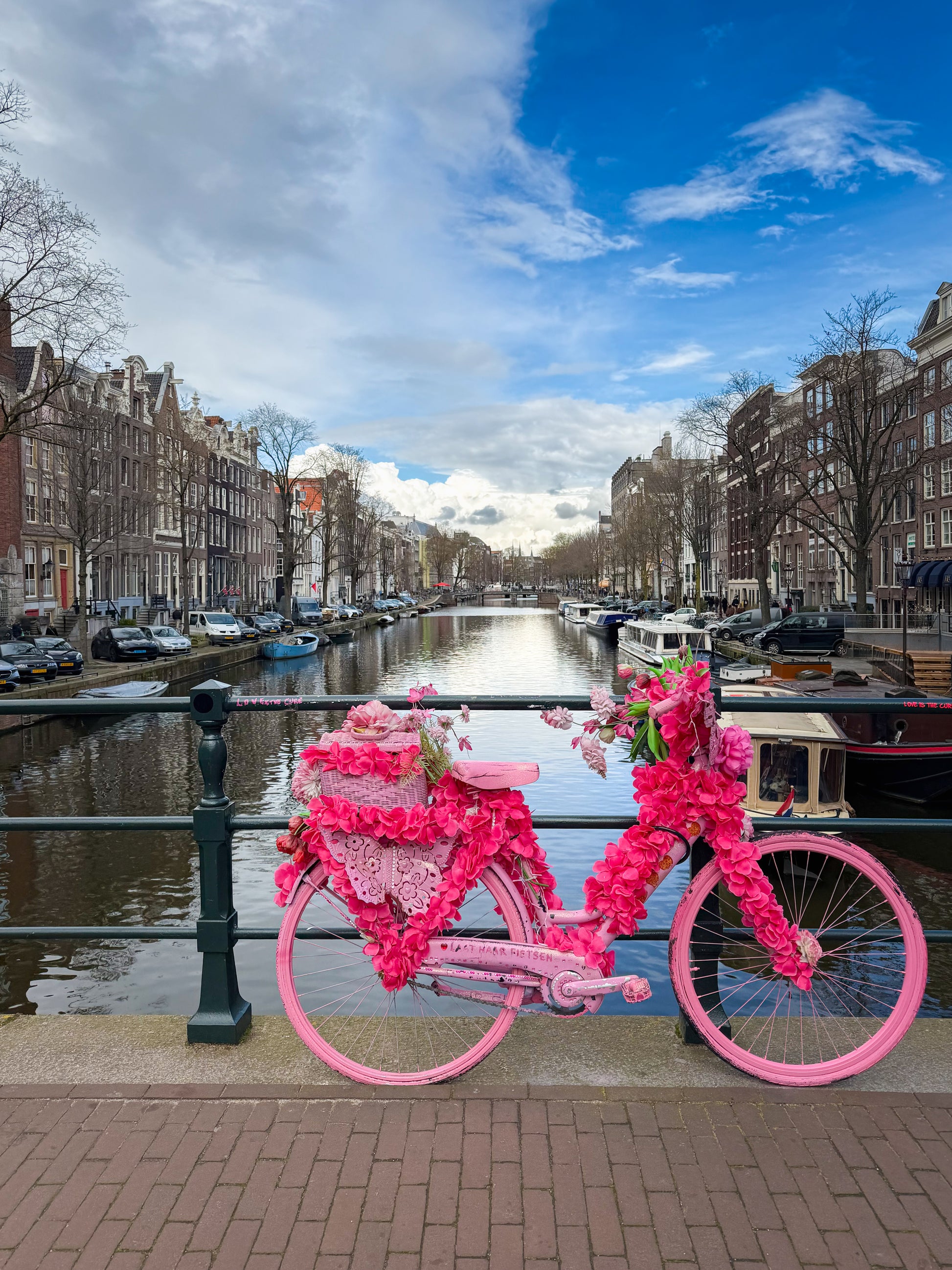 Flower-covered pink bicycle on an Amsterdam canal bridge with historic buildings, boats, water, and city scenery in the background.