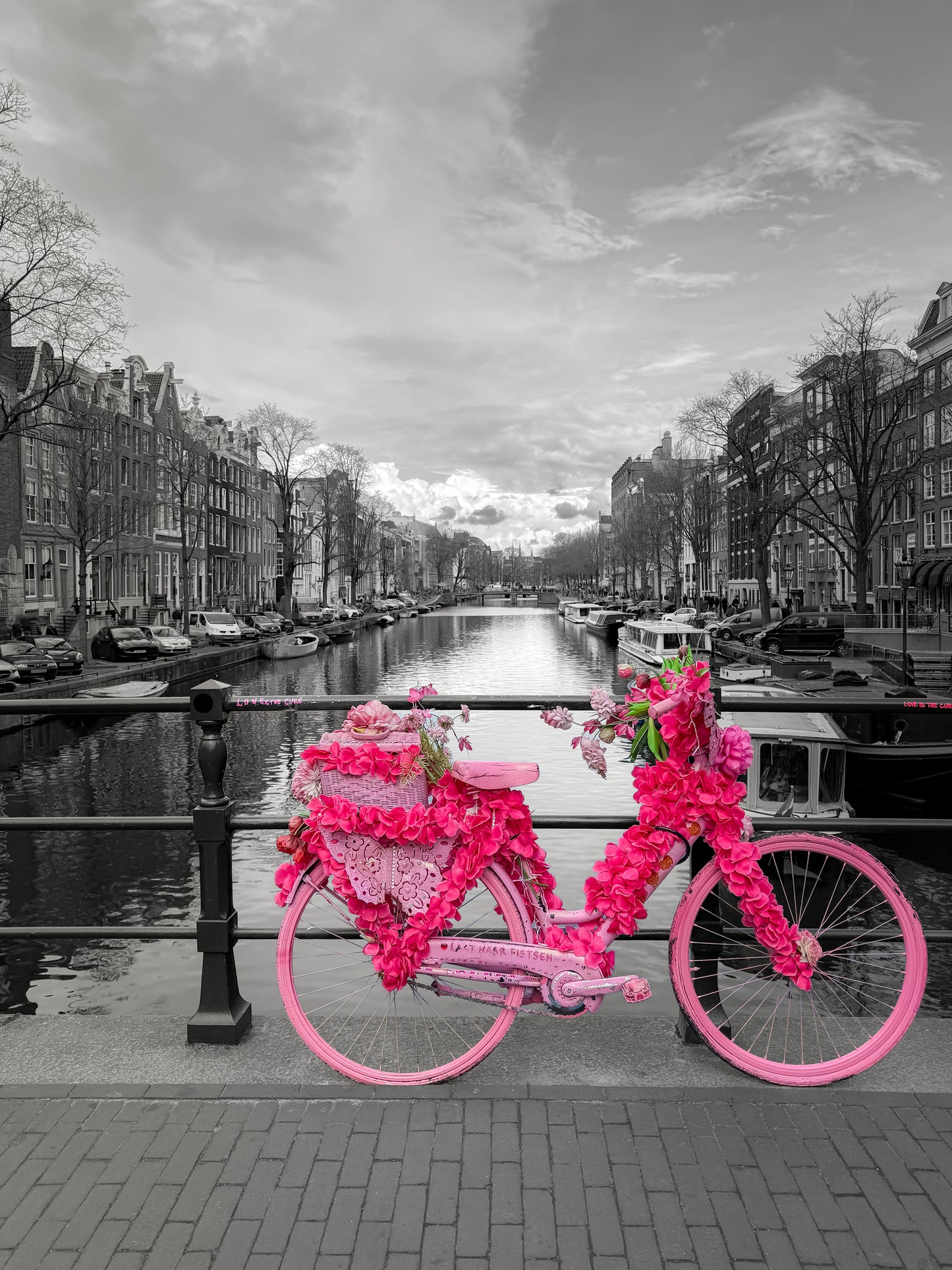 Flower-covered pink bicycle on an Amsterdam canal bridge with historic buildings, boats, water, and city scenery in the background. Printed in B&W with pink detail.