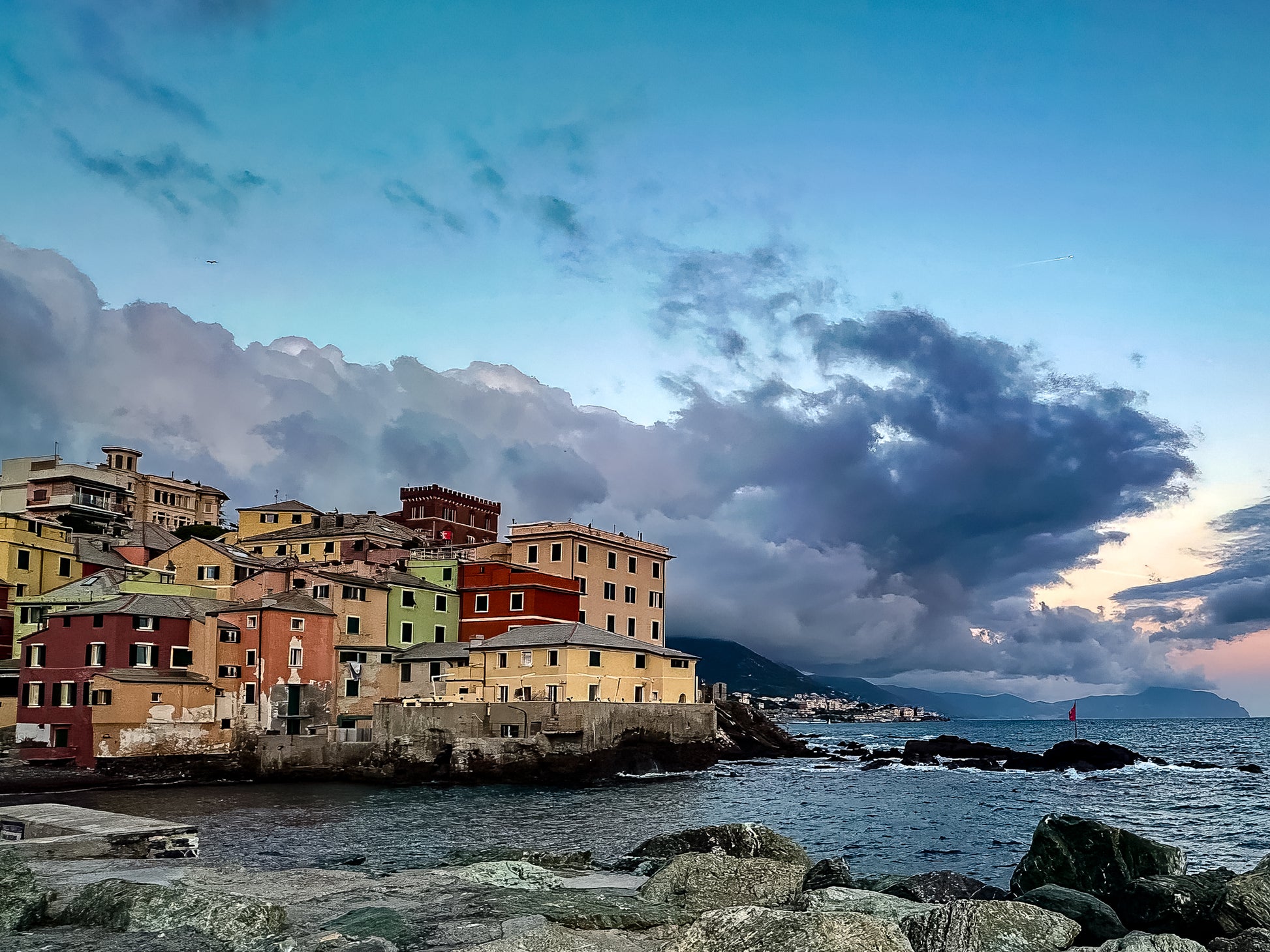 Colorful buildings along the Ligurian Sea in Genoa, Italy, with rocky shoreline, deep blue water, distant mountains, and dramatic evening clouds.