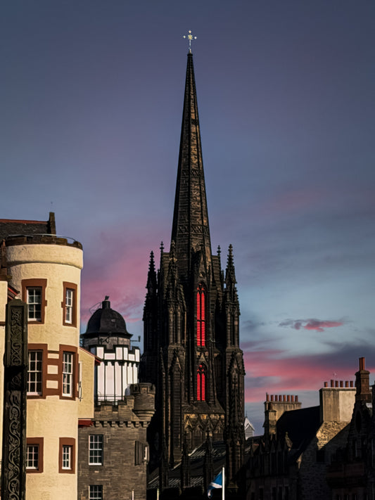 Dark church spire rising above historic buildings in Edinburgh, Scotland, with glowing red windows and a soft evening sky.