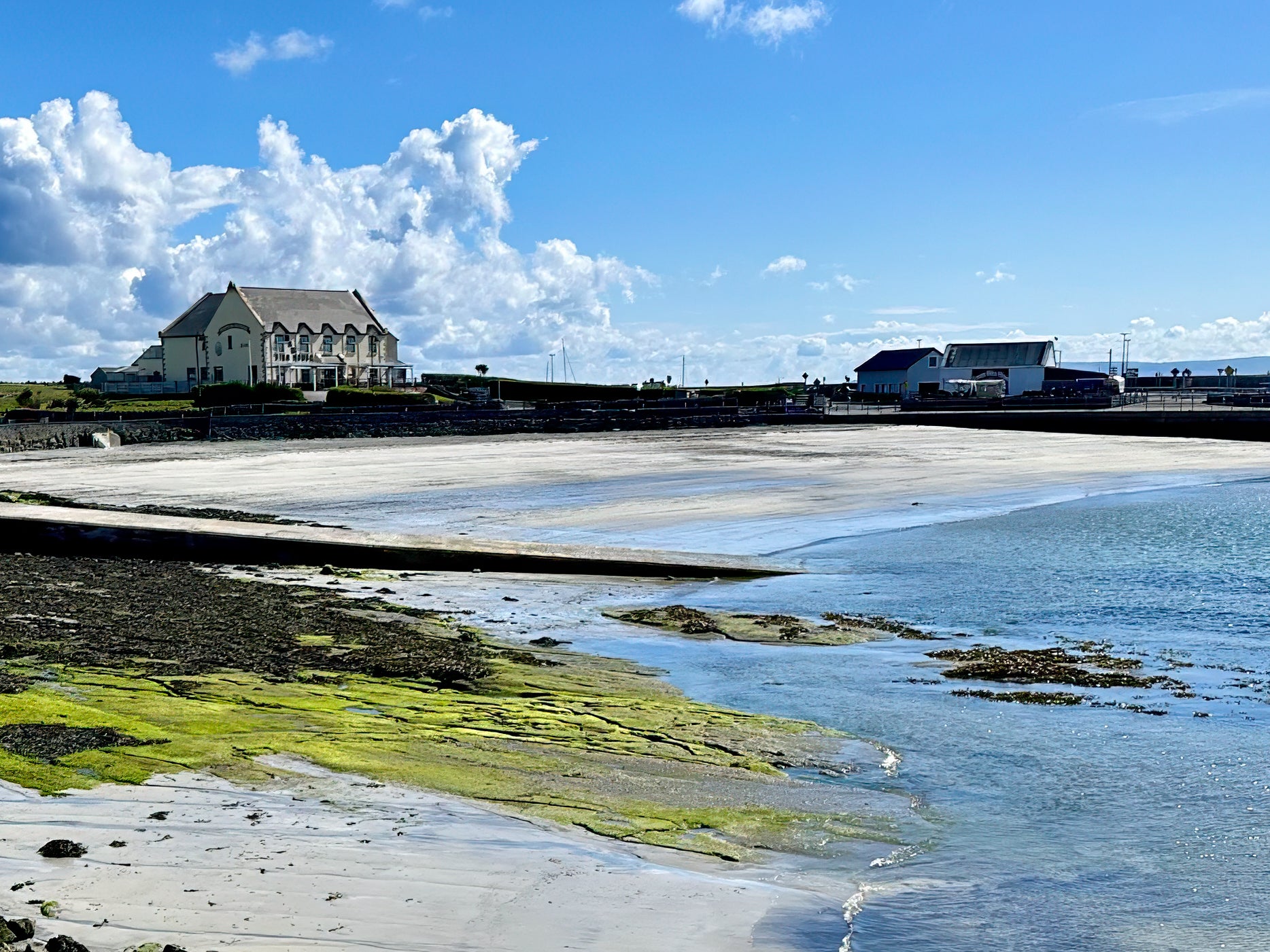 Ireland coastal landscape at low tide with blue water, soft sand, green shoreline textures, buildings, and open sky.
