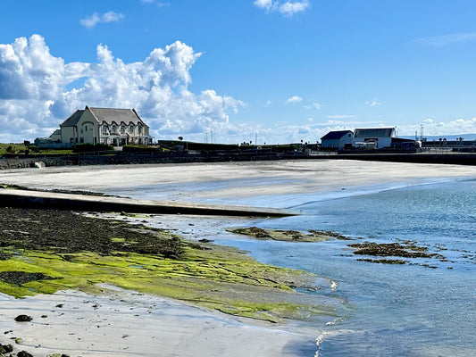 Ireland coastal landscape at low tide with blue water, soft sand, green shoreline textures, buildings, and open sky.