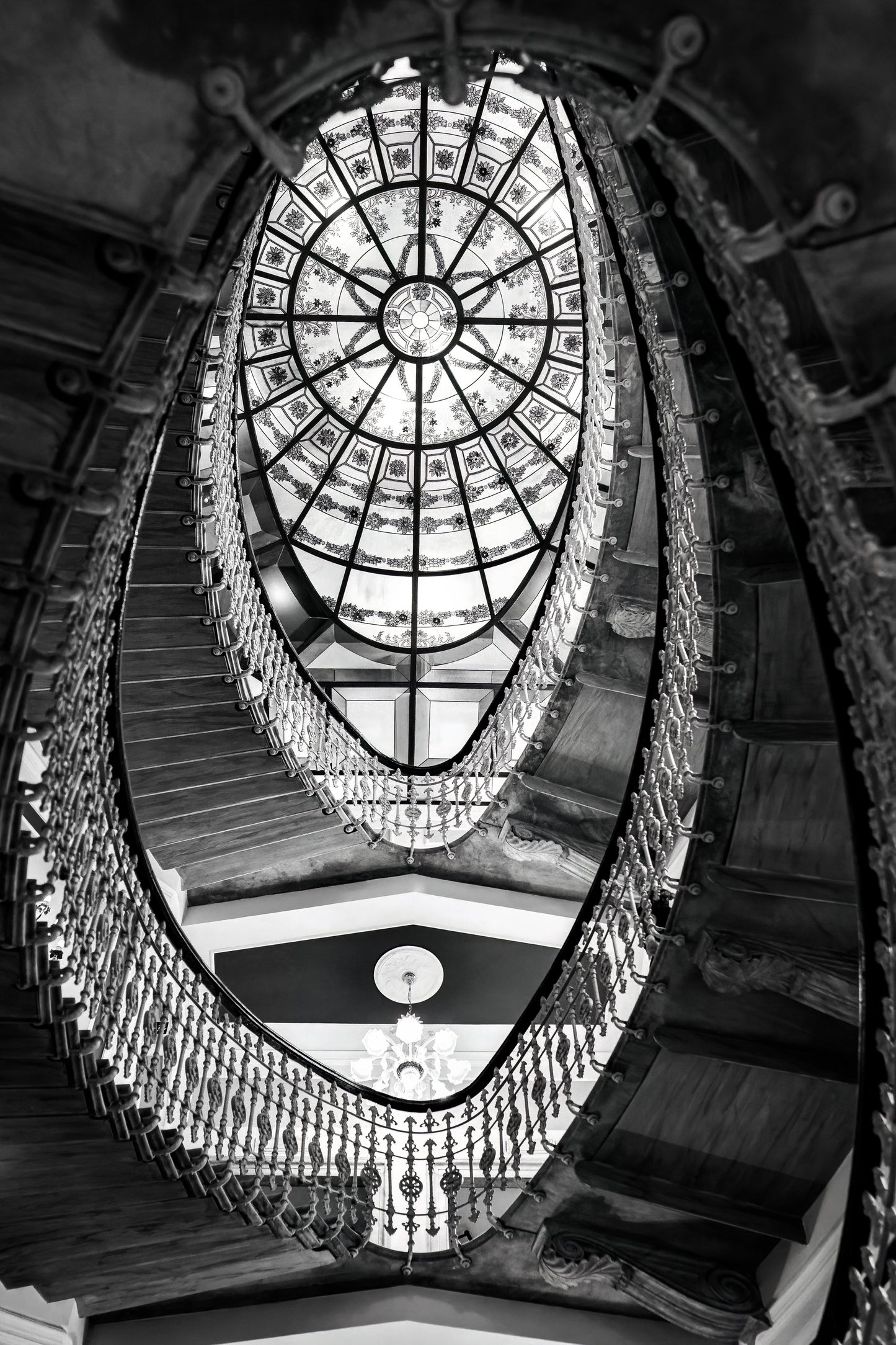 Black and white photograph of the elliptical staircase inside Hotel Bristol Palace in Genoa, Italy, with ornate railing and glass ceiling above.
