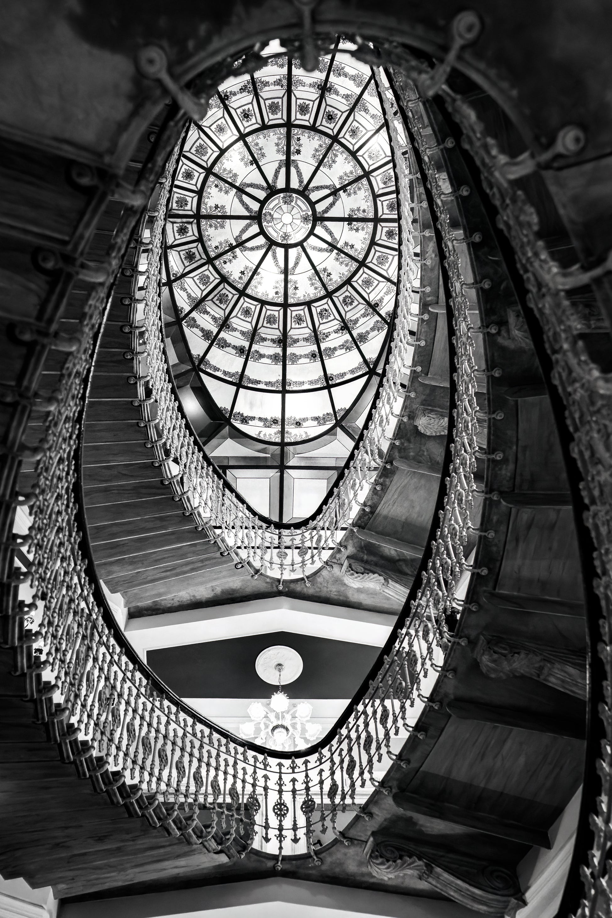 Black and white photograph of the elliptical staircase inside Hotel Bristol Palace in Genoa, Italy, with ornate railing and glass ceiling above.