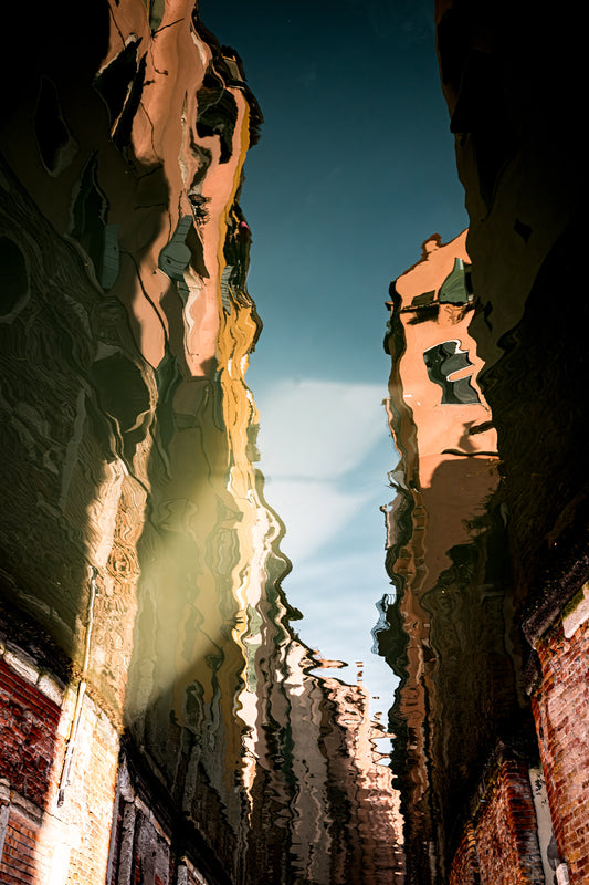 Abstract reflection of Venice buildings in canal water with warm tones, deep shadows, brick walls, and soft painterly movement.